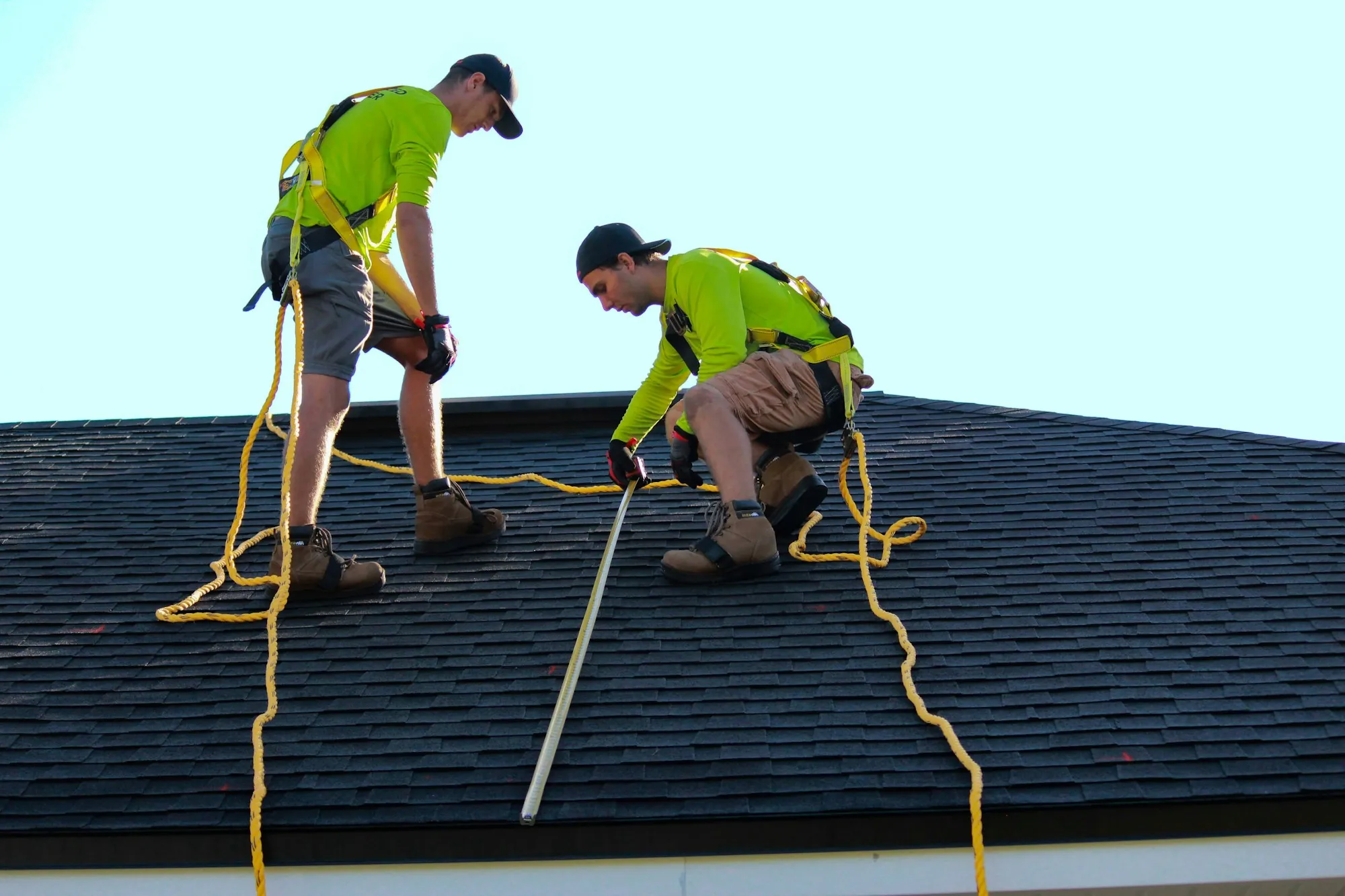 Two roofers in safety harnesses measuring a dark shingle roof during a roof replacement in Brookhaven